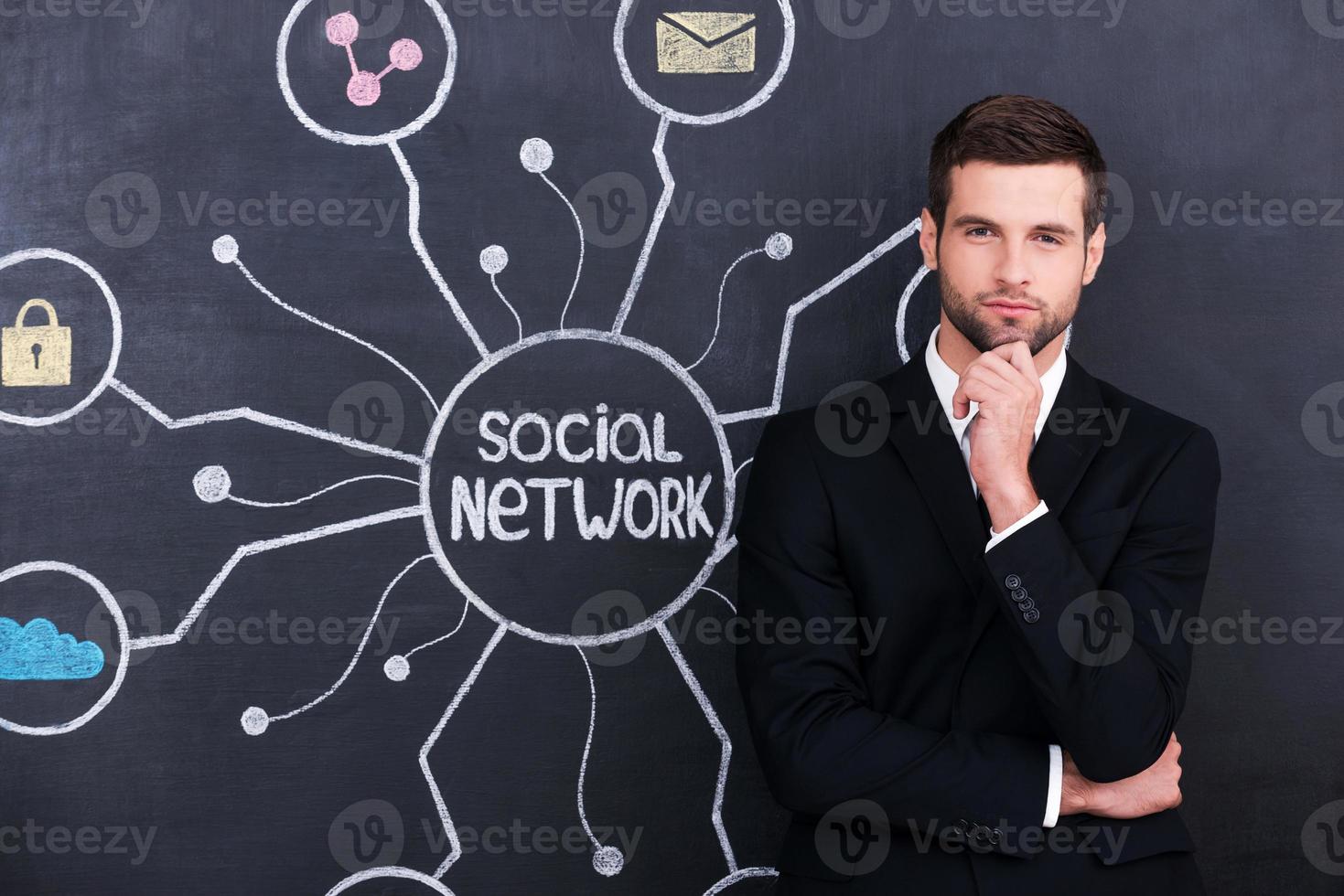 Being social active person. Handsome young man holding hand on chin and looking at camera while standing against social network chalk drawing on blackboard photo