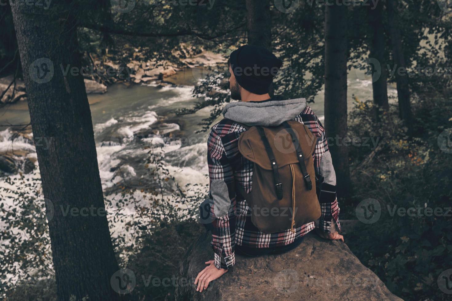 Enjoy the silence. Rear top view of young modern man with backpack sitting in the woods before the river while spending carefree time in mountains photo