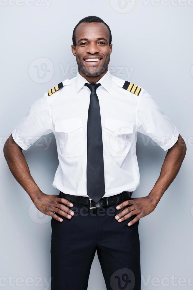 Confident pilot. Confident African pilot in uniform holding hand on hip and smiling while standing against grey background photo