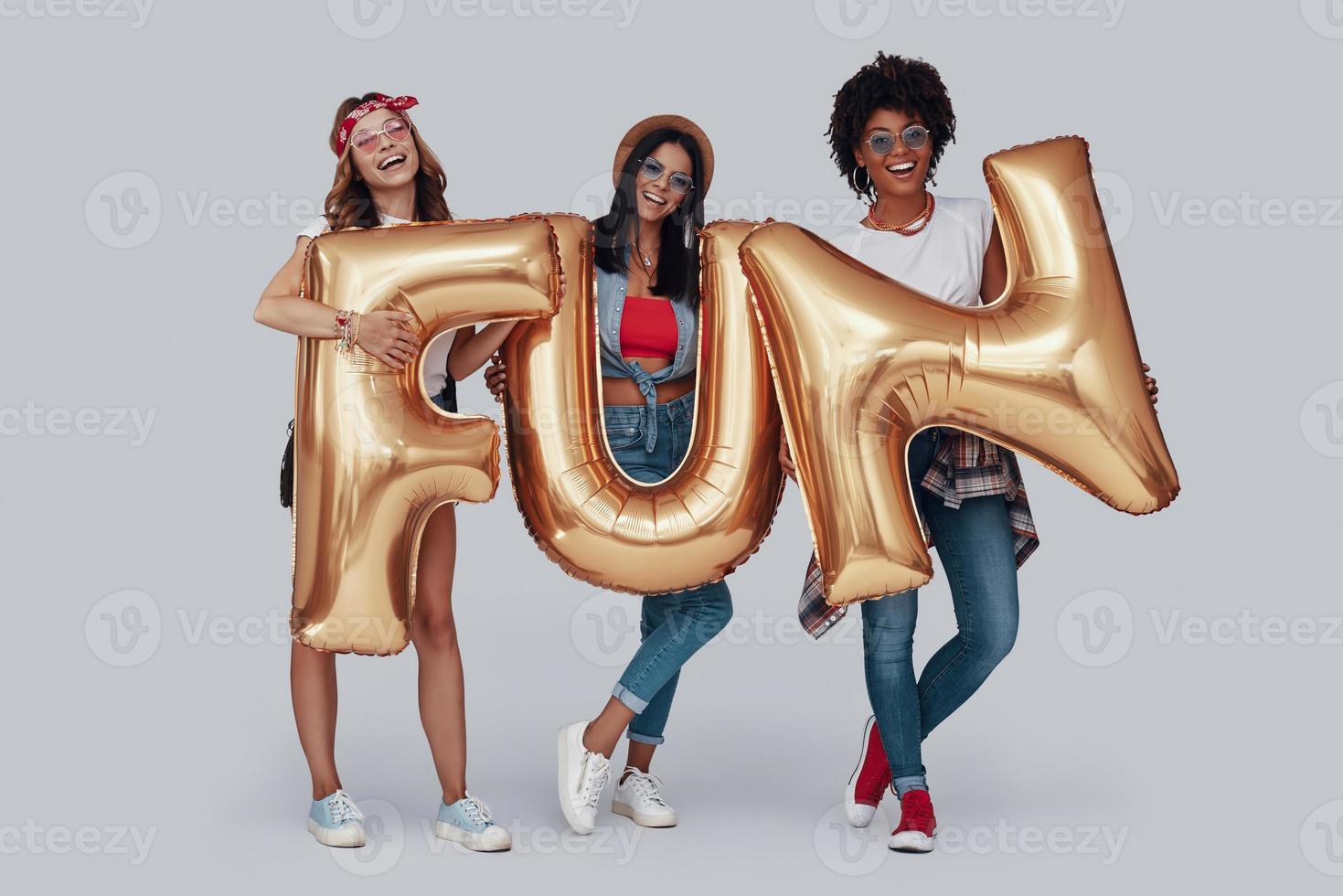 Full length of three attractive young women carrying balloons and smiling while standing against grey background photo