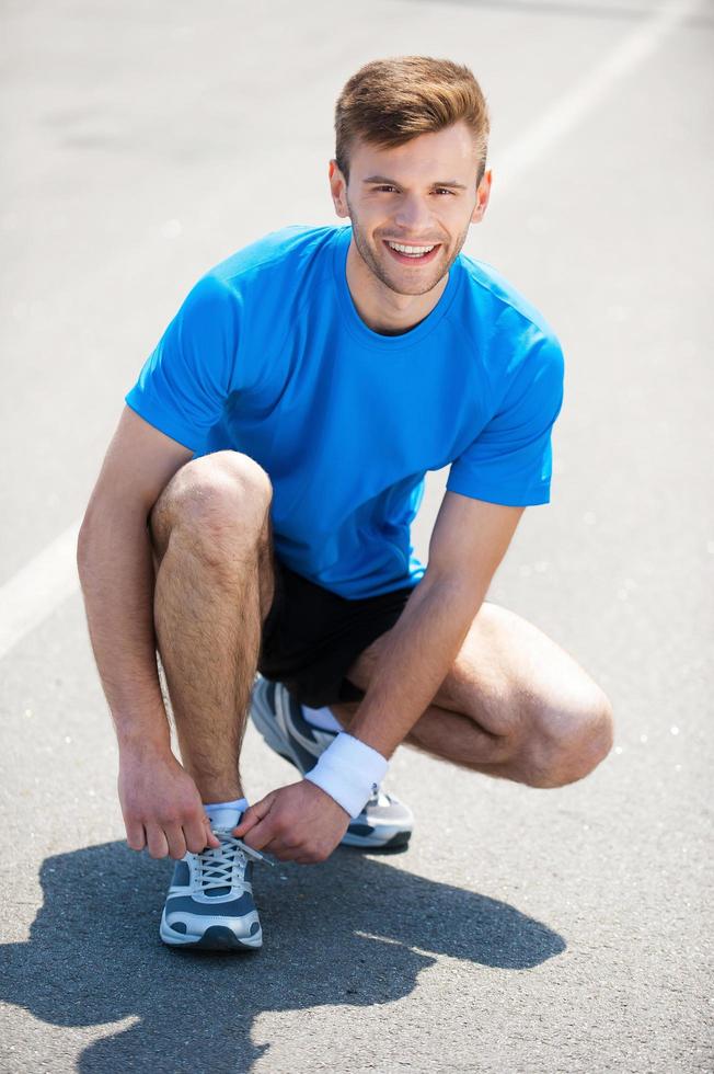 Getting ready to jogging. Top view of man tying shoelaces on sports shoe and smiling while standing outdoors photo