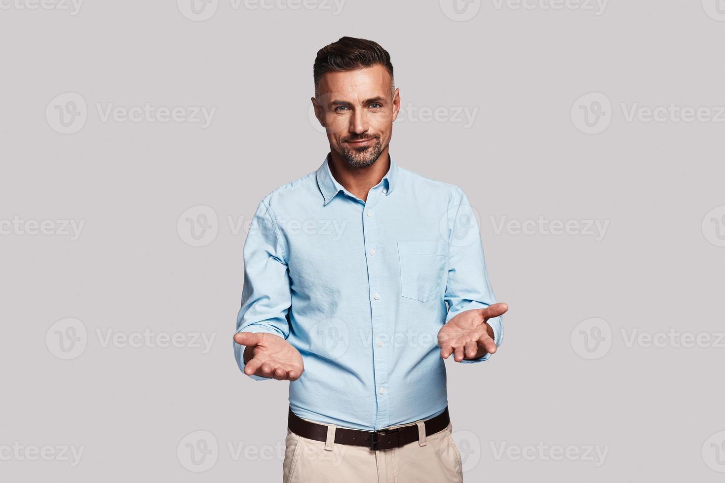 Welcome Good looking young man keeping arms outstretched and looking at camera while standing against grey background photo