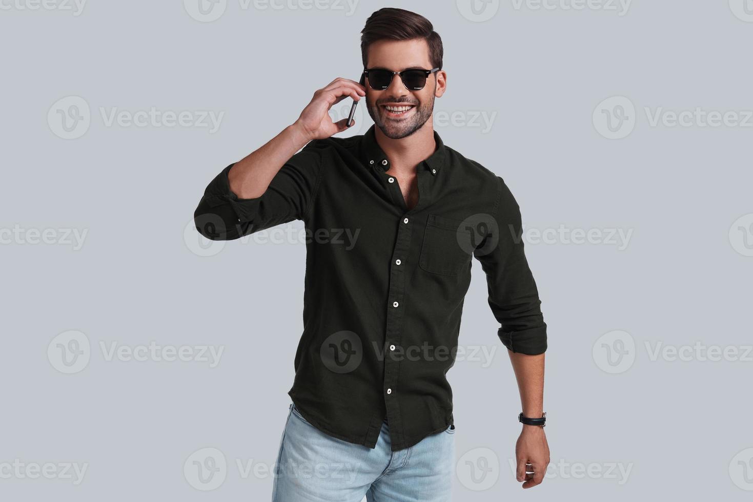 Confident and handsome.  Good looking young man in smart casual wear talking on his smart phone and smiling while standing against grey background photo