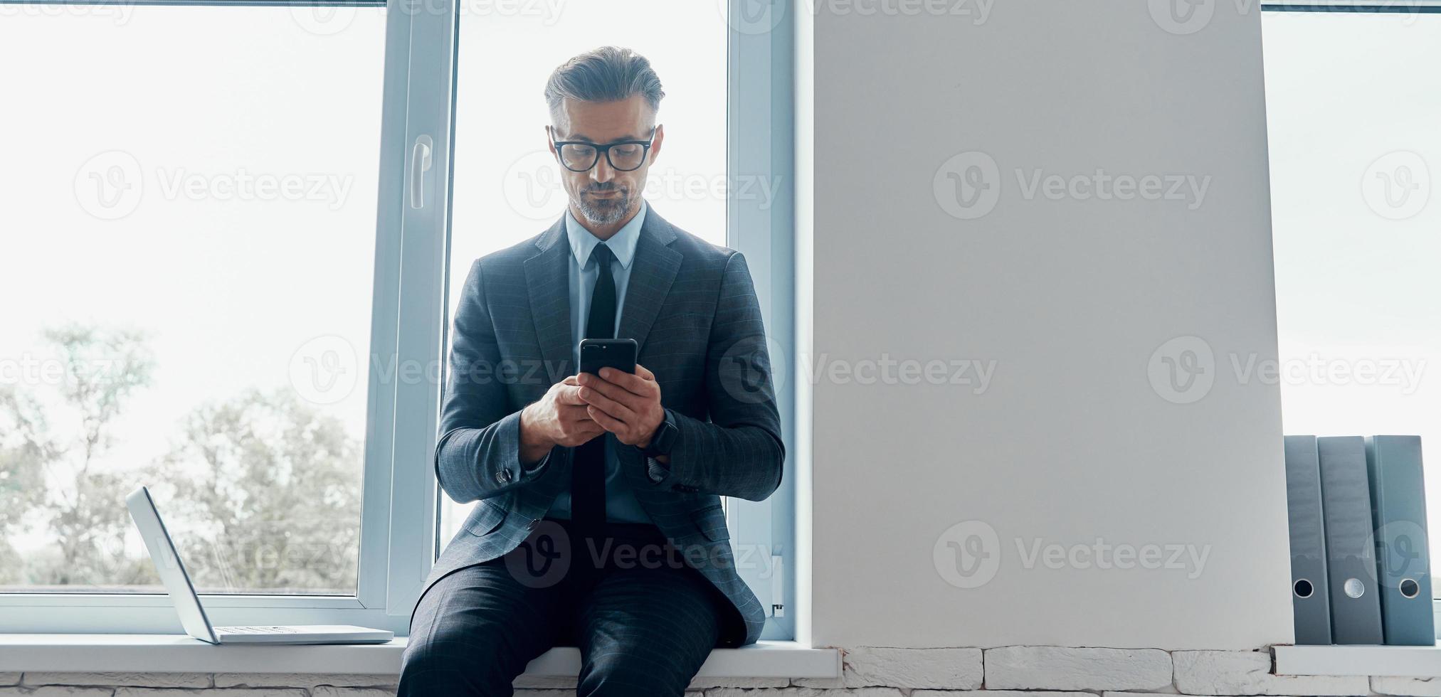 Confident mature man in formalwear using smart phone while sitting on the window sill in office photo