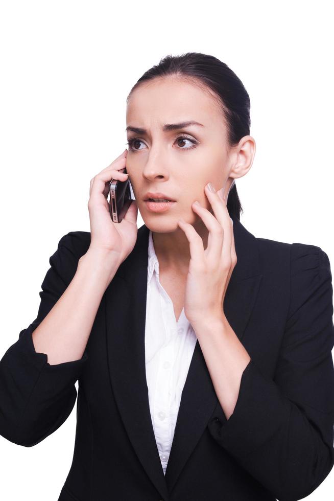 Bad news. Frustrated young woman in formalwear talking on the mobile phone and touching her face with hand while standing isolated on white photo