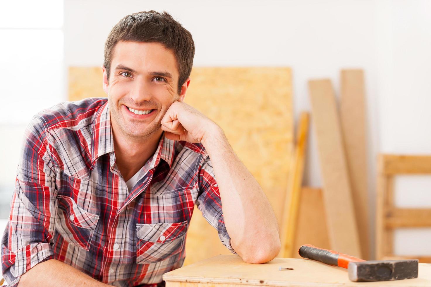 tomando tiempo para relajarse. apuesto joven manitas sonriendo mientras se sienta en el taller y se apoya en la plataforma de madera foto