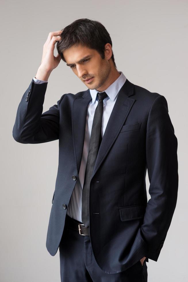 Lost in business thoughts. Thoughtful young man in formalwear holding hand in hair and looking away while standing against grey background photo