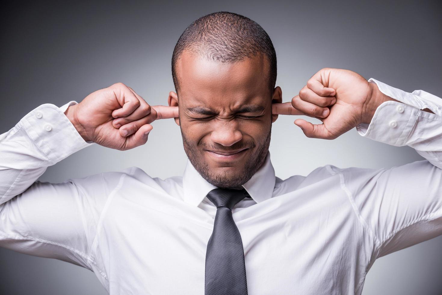 Too loud sound. Young African man in shirt and tie covering ears with hand while standing against grey background photo