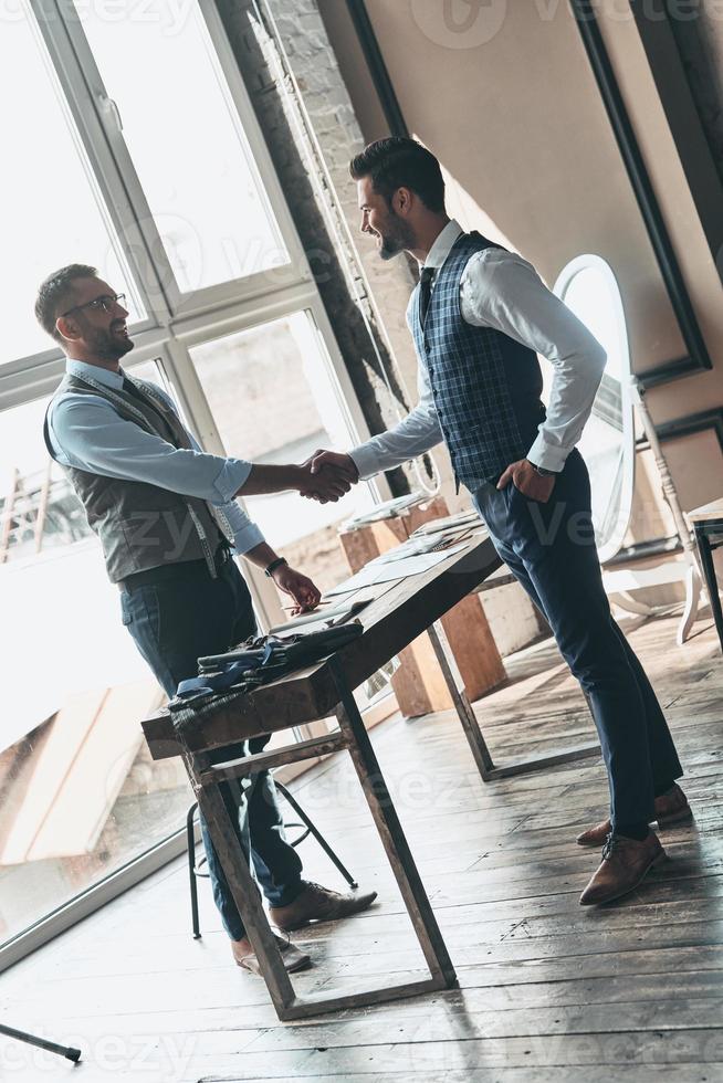 Greeting new client. Full length of two young fashionable men shaking hands and smiling while standing in workshop photo