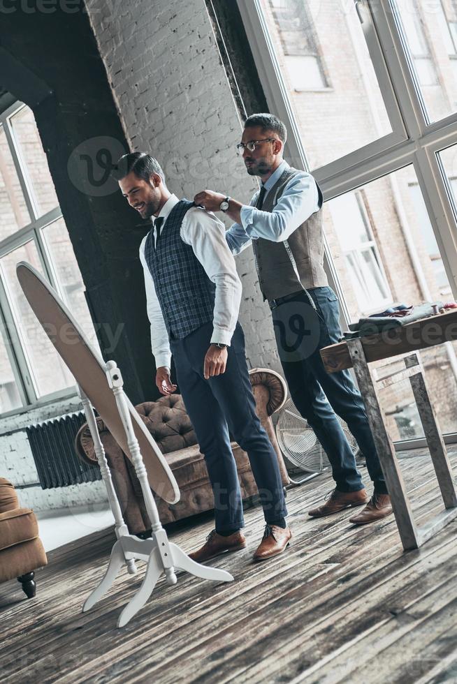 Fits perfect. Full length of young fashionable designer helping his client to get dressed while standing in his workshop photo