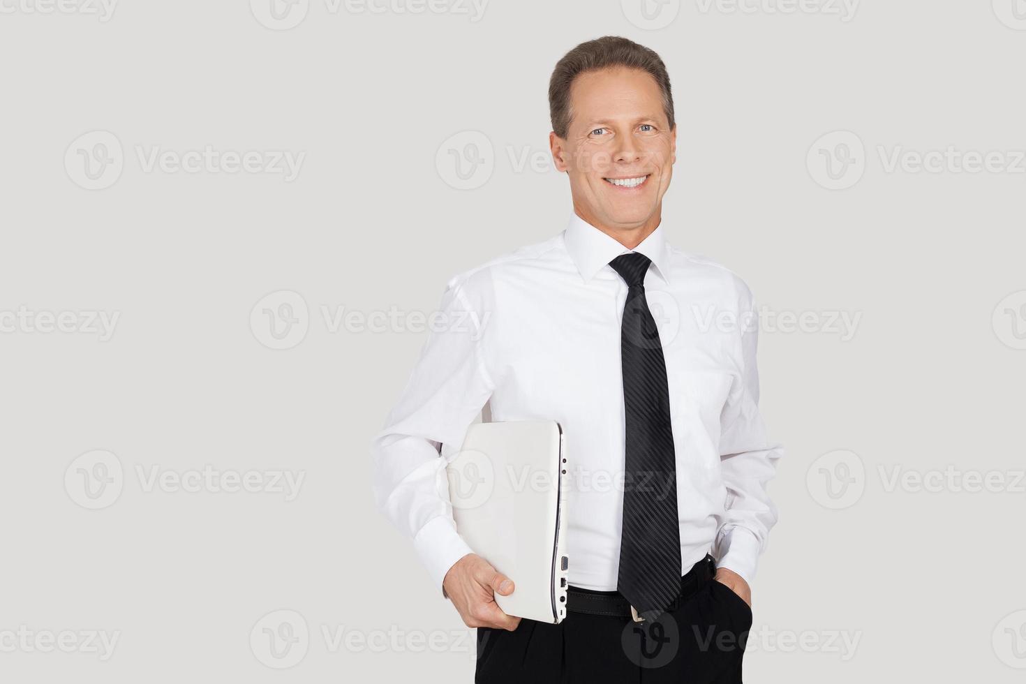 Businessman with laptop. Cheerful senior man in formalwear holding laptop and smiling while standing against grey background photo