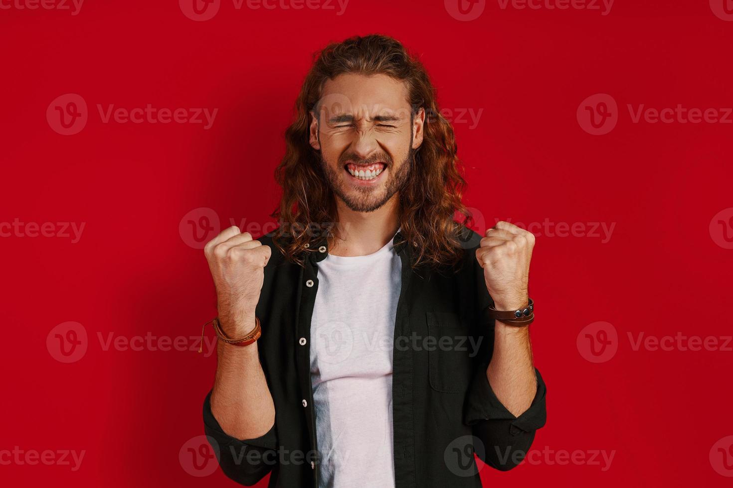 Cheerful young man in casual clothing smiling and gesturing while standing against red background photo