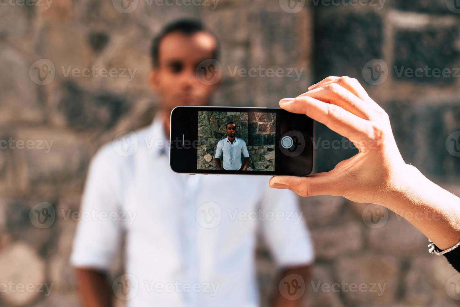 Taking a picture. Female hand holding smart phone and taking picture of young African man standing outdoors photo