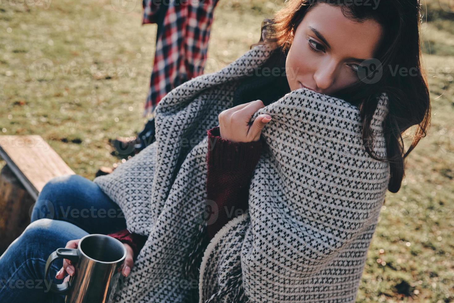 Feeling free and relaxed. Beautiful young woman covered with blanket holding a mug and looking away while spending time by the campfire with her boyfriend photo