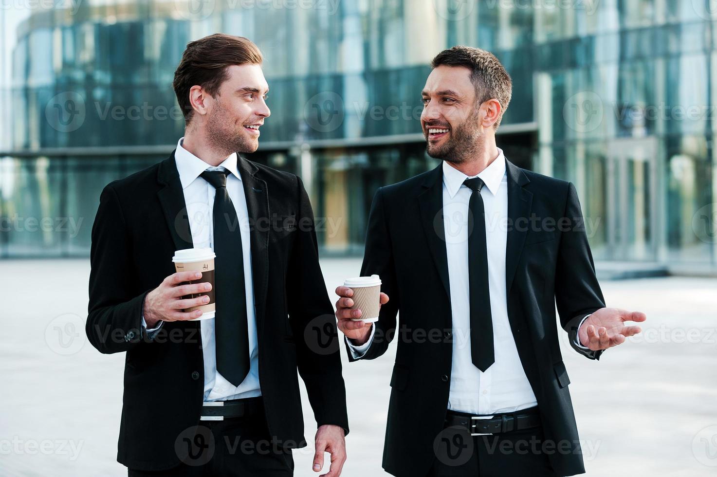Colleagues and friends. Two cheerful businessmen holding cups on coffee and talking to each other while walking outdoors photo