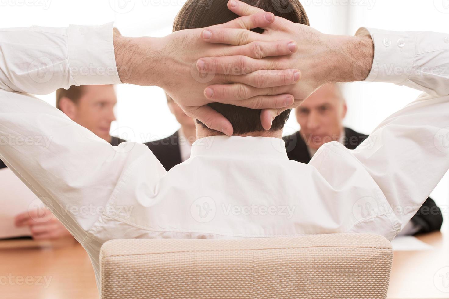 Feeling confident. Rear view of man in white shirt holding hands behind head while three colleagues sitting in front of him photo