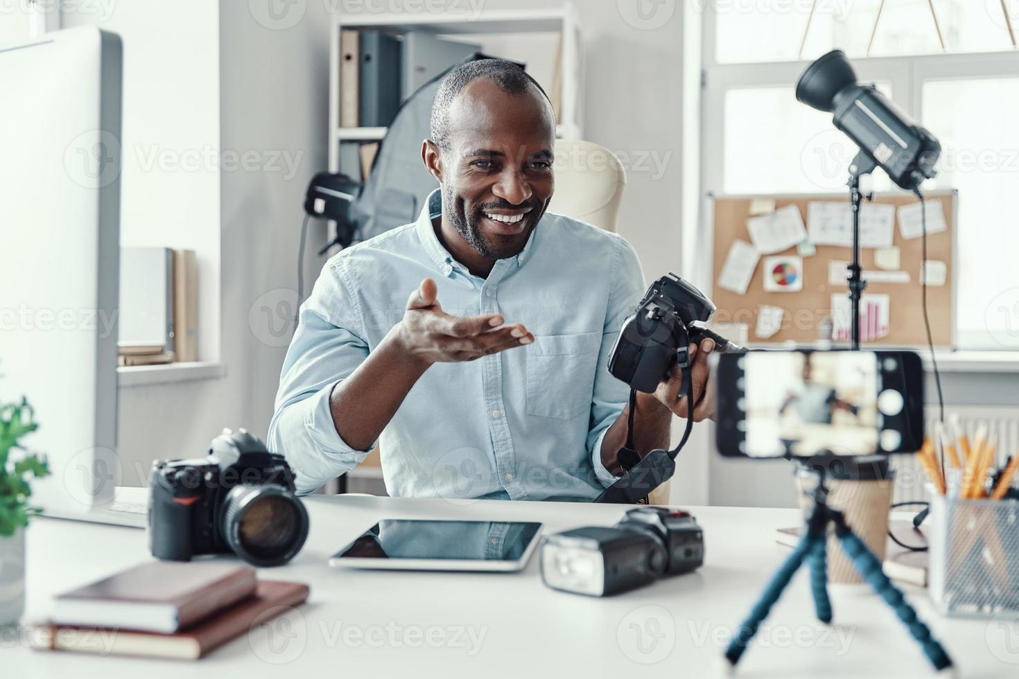 Charming young African man in shirt showing digital camera and telling something while making social media video photo