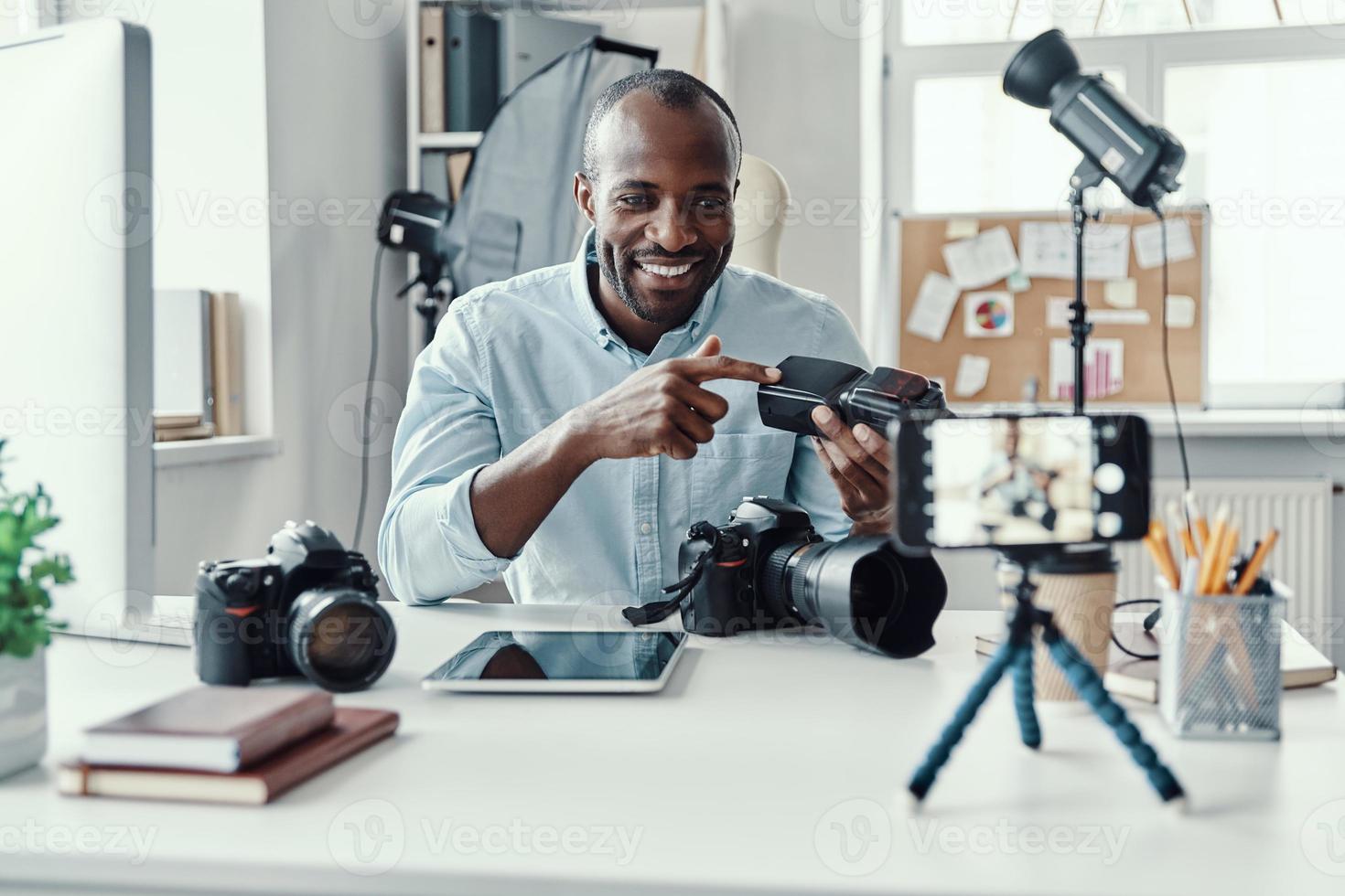 Charming young African man in shirt showing digital camera and telling something while making social media video photo