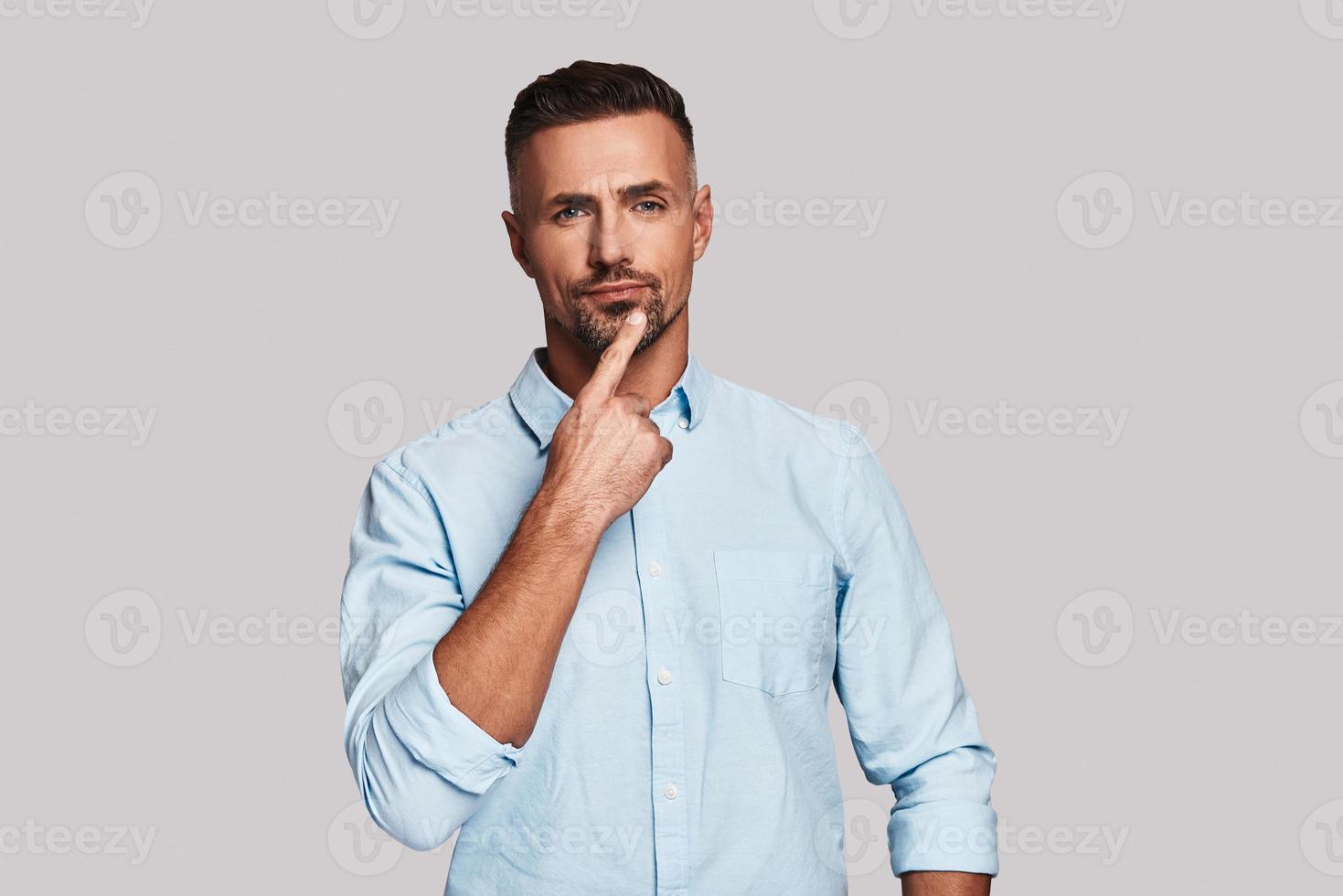 Uncertainty. Thoughtful young man keeping hand on chin and looking at camera while standing against grey background photo