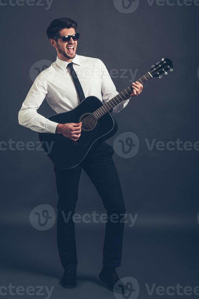 Rock or business star Full length of handsome well-dressed young man in sunglasses playing guitar and keeping mouth open while standing against grey background photo