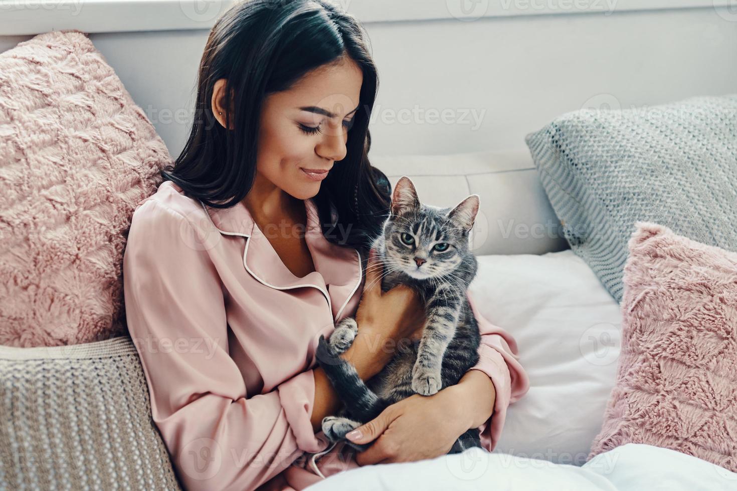 Beautiful young woman in pajamas smiling and embracing domestic cat while resting in bed at home photo