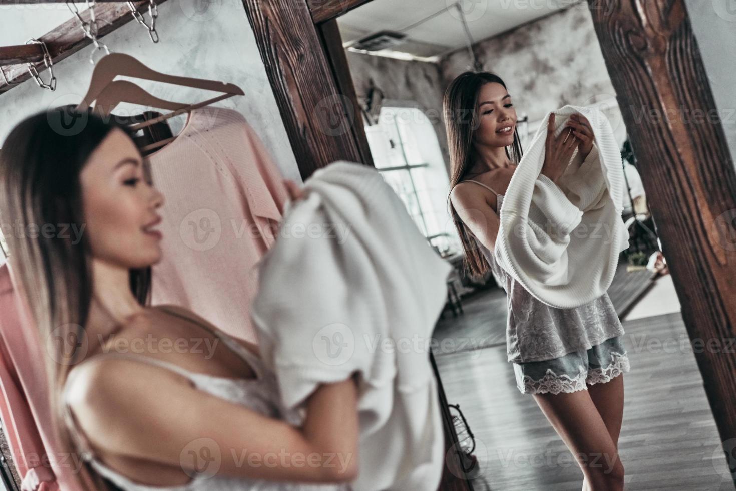 Extremely beautiful. Attractive young woman taking on the blouse and smiling while standing in front of the mirror at home photo