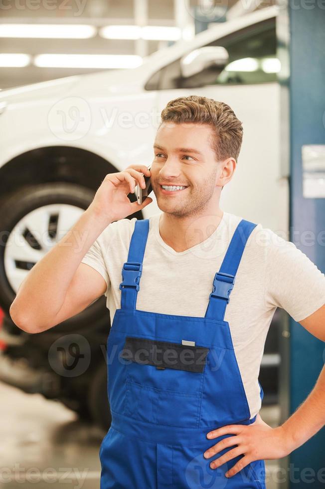 Your car is like a new Handsome young man talking on the mobile phone and smiling while standing in workshop with car in the background photo