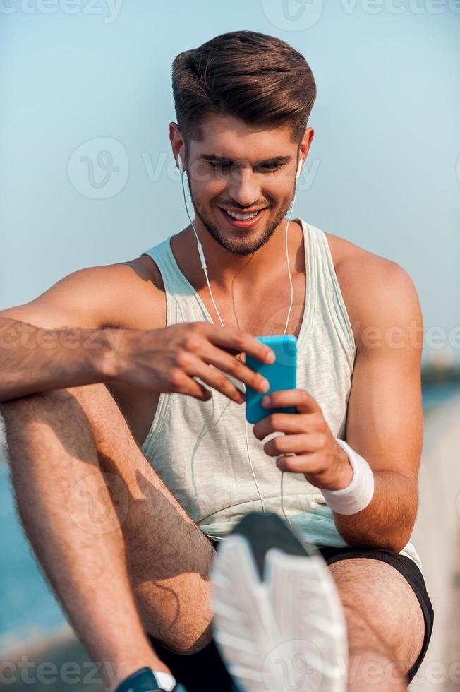 la elección de la música para el entrenamiento. feliz joven musculoso con auriculares mirando este reproductor de mp3 y sonriendo mientras se sienta en el parapeto foto