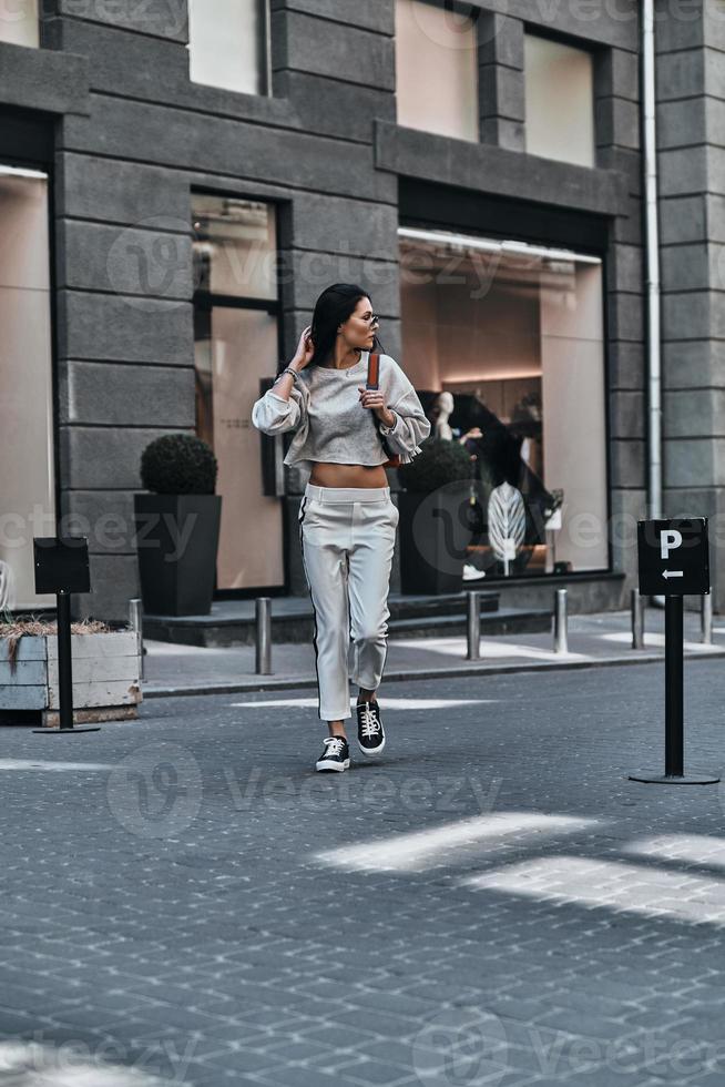 City style. Beautiful young woman keeping hand in hair and looking away while walking outdoors photo