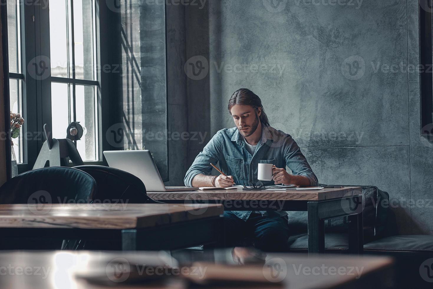 Taking notes. Thoughtful young man in smart casual wear writing something and holding a cup while sitting at the office desk photo