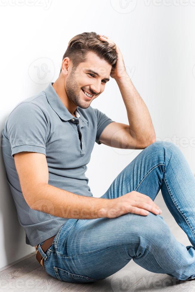 alegre guapo. un joven guapo cogido de la mano en el pelo y sonriendo mientras se sienta en el suelo y se apoya en la pared foto