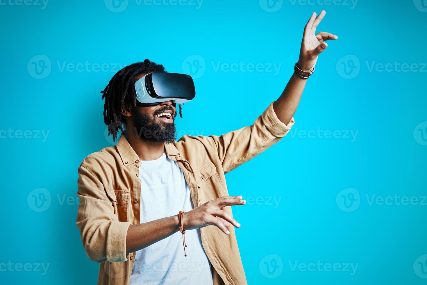 alegre joven africano con gafas de realidad virtual mientras está de pie contra el fondo azul foto