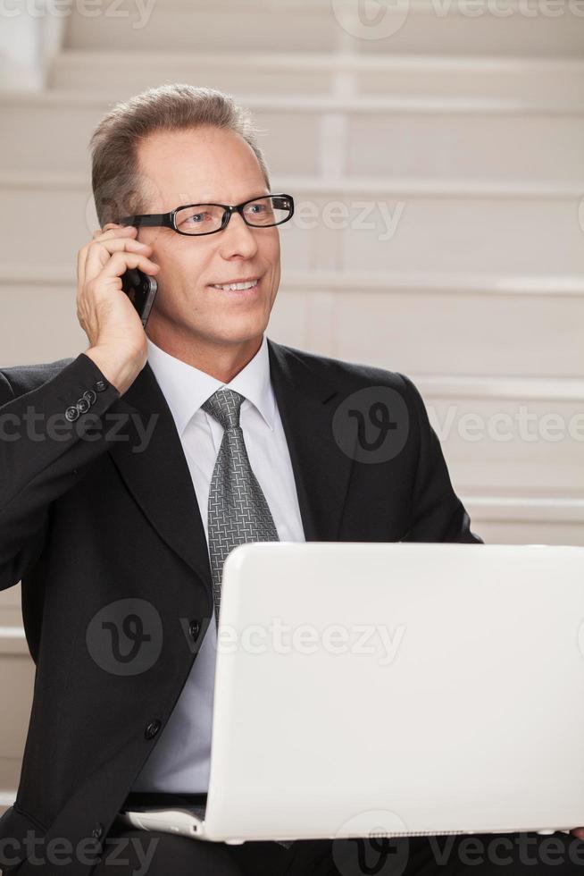 He is always in touch. Cheerful mature man in formalwear talking on the mobile phone and using computer while sitting on staircase photo