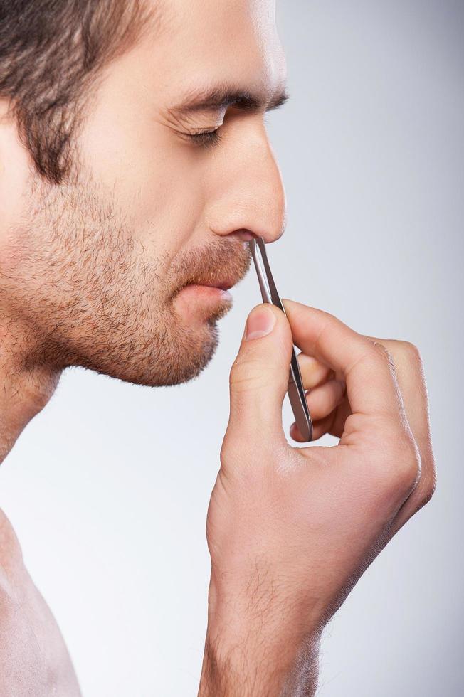 Man tweezing hair from nose. Side view of young man tweezing hair from nose and keeping eyes closed while standing against grey background photo