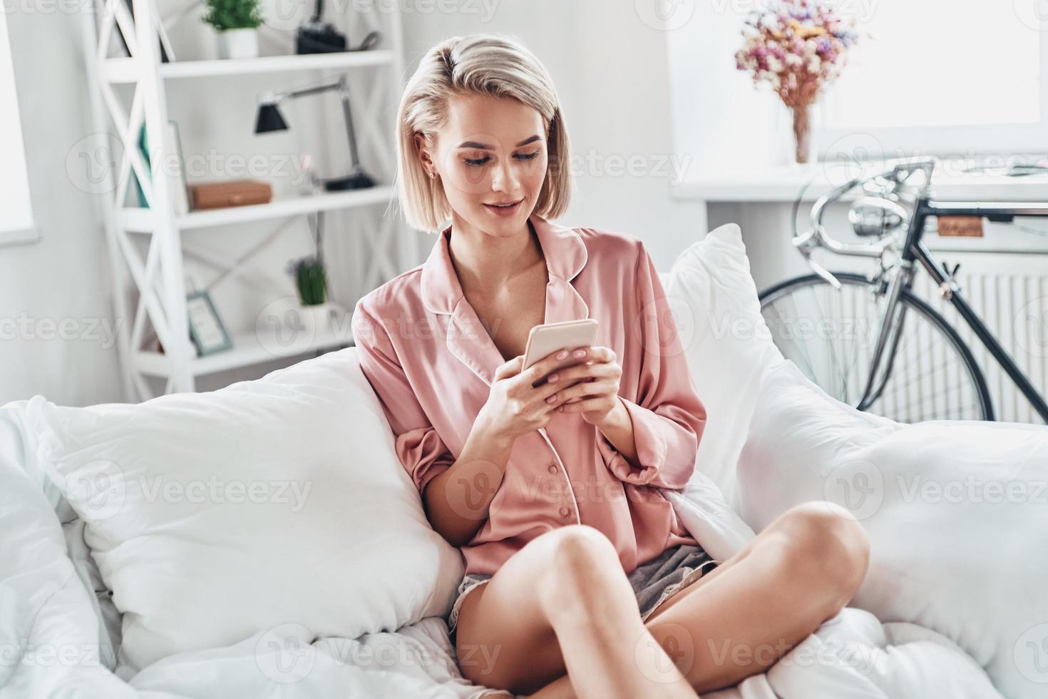 Feeling lazy. Attractive young woman using smart phone and smiling while sitting on bed at home photo