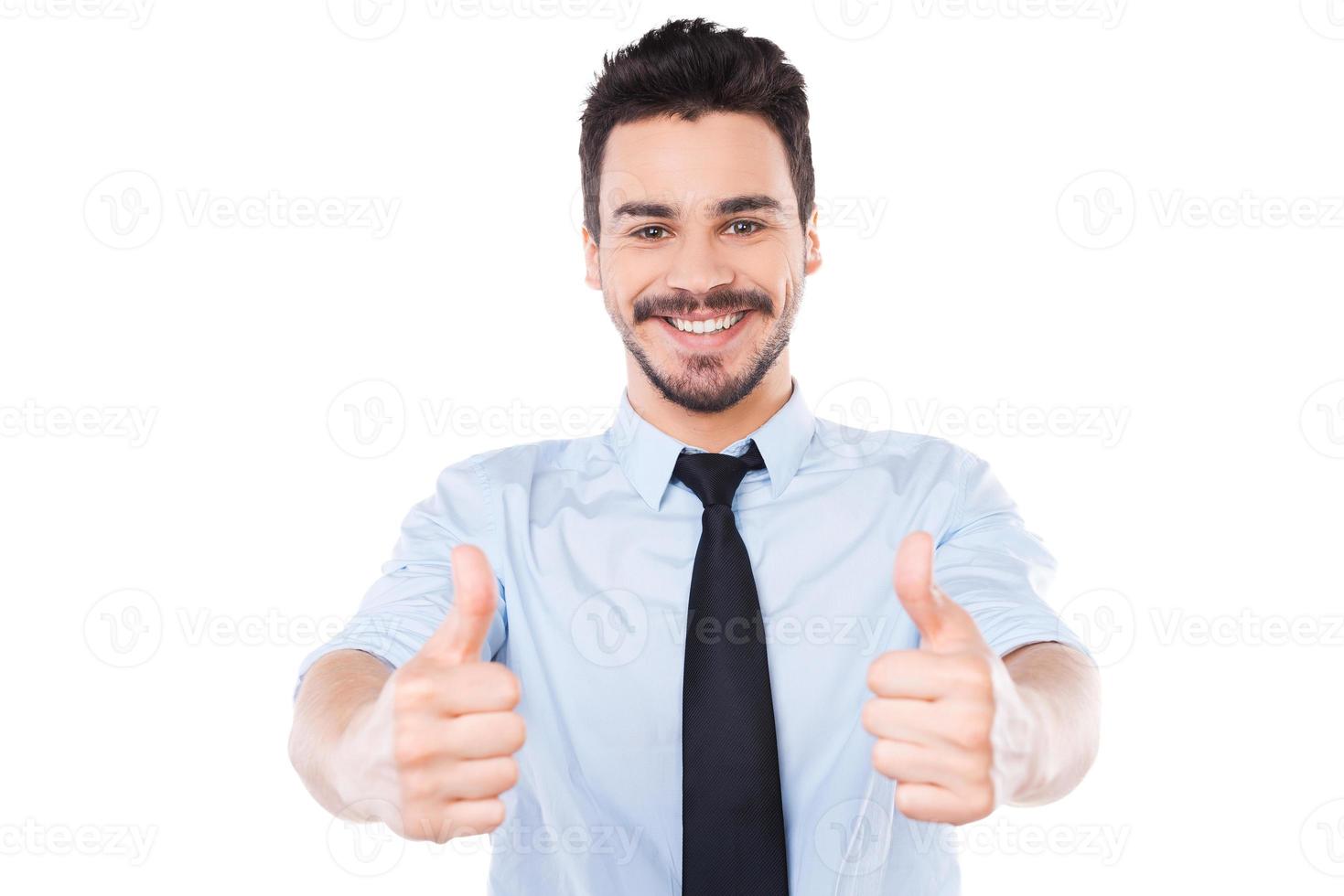 I say yes Confident young man in shirt and tie looking at camera and showing his thumbs up and smiling while standing against white background photo