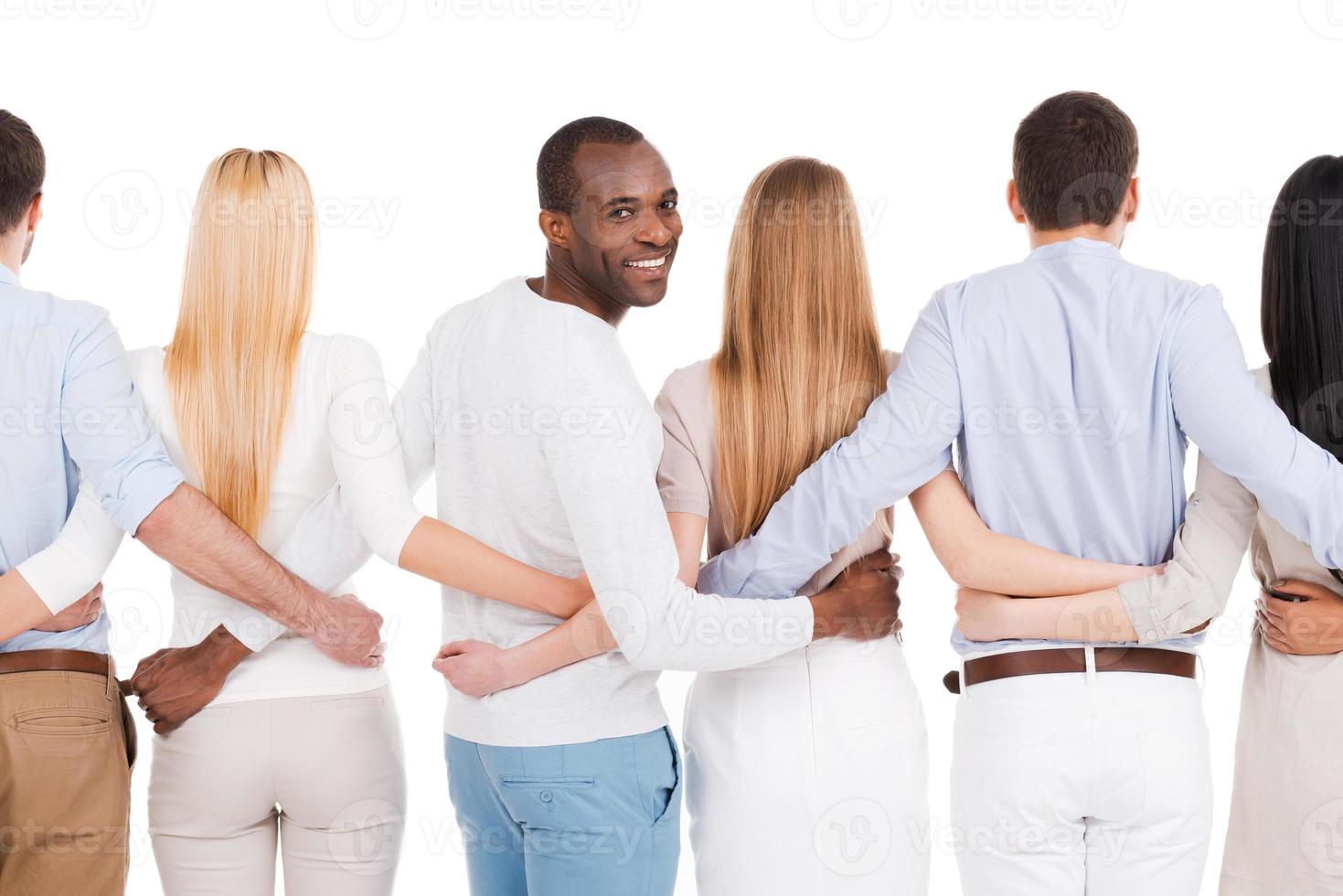 Together we are stronger Rear view of group of diverse people bonding to each other and standing against white background while one African man looking over shoulder and smiling photo