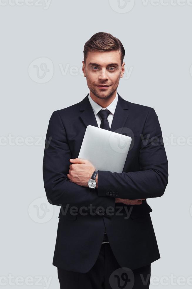 Young and successful. Handsome young man carrying digital tablet and looking at camera with smile while standing against grey background photo