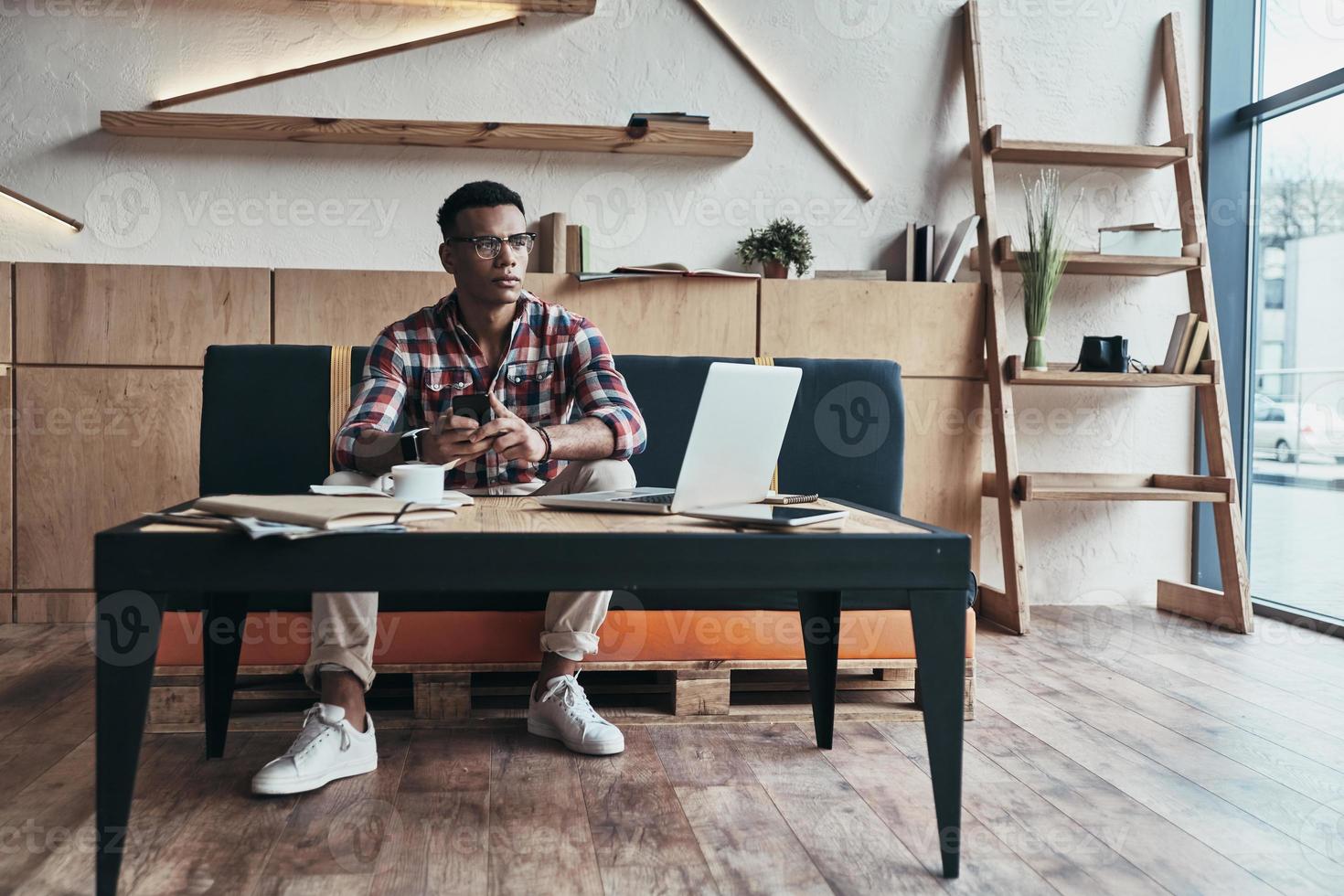 Searching for best answer. Thoughtful young man using his smart phone and looking away while sitting in the creative working space photo