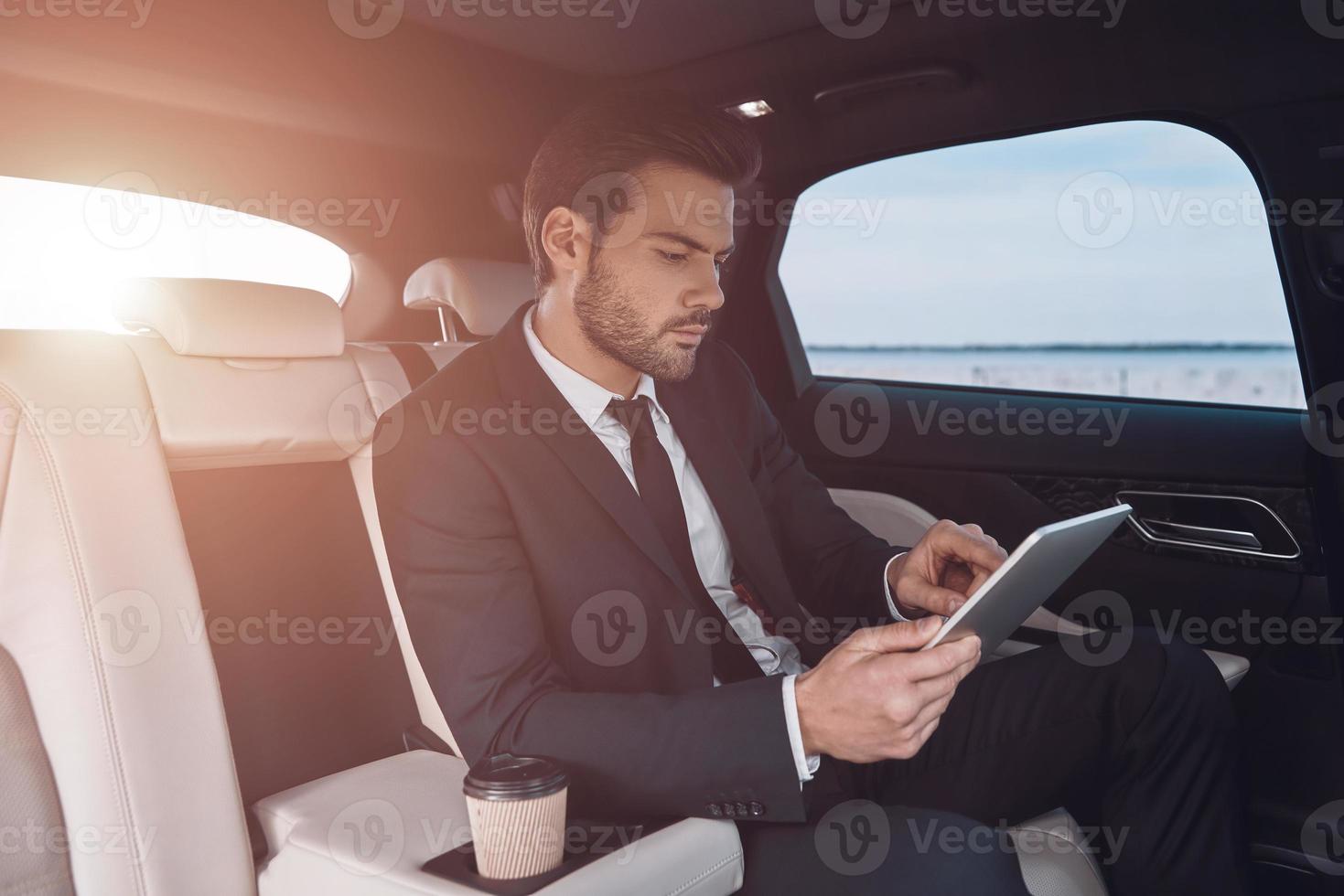 teniendo en cuenta el siguiente paso. un joven apuesto con traje completo que trabaja con una tableta digital mientras está sentado en el auto foto