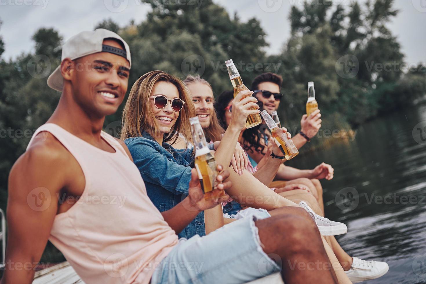 Real friendship. Group of happy young people in casual wear smiling and looking at camera while sitting on the pier photo