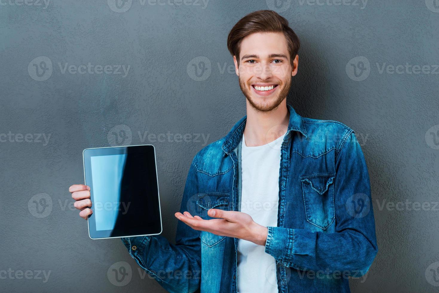 Your advertisement on his digital tablet. Smiling young man holding digital tablet and pointing at it while standing against grey background photo