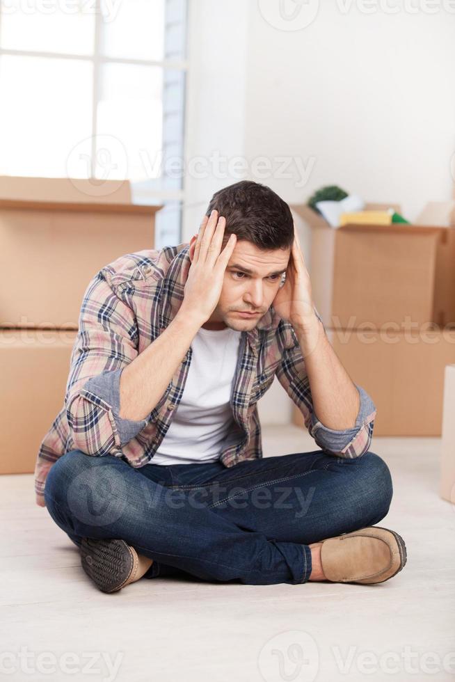 So many boxes to unpack. Depressed young man sitting on the floor and holding head in hands while cardboard boxes laying on the background photo