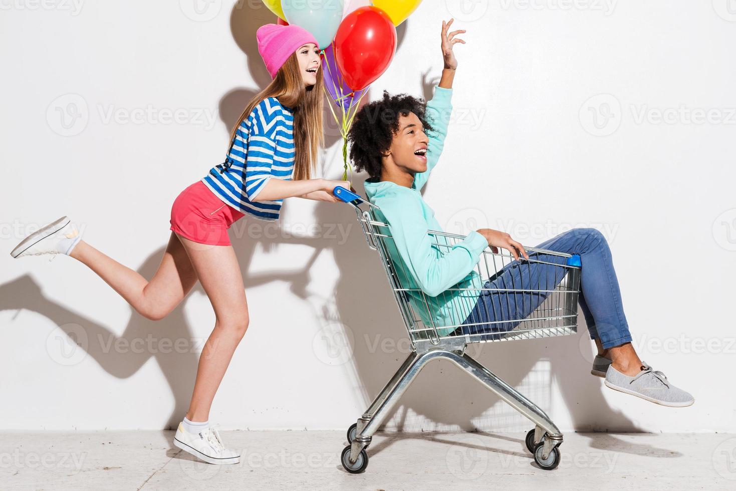 pasar un buen rato juntos. mujeres jóvenes felices que llevan a su novio en un carrito de compras y sonríen mientras corren contra un fondo gris foto