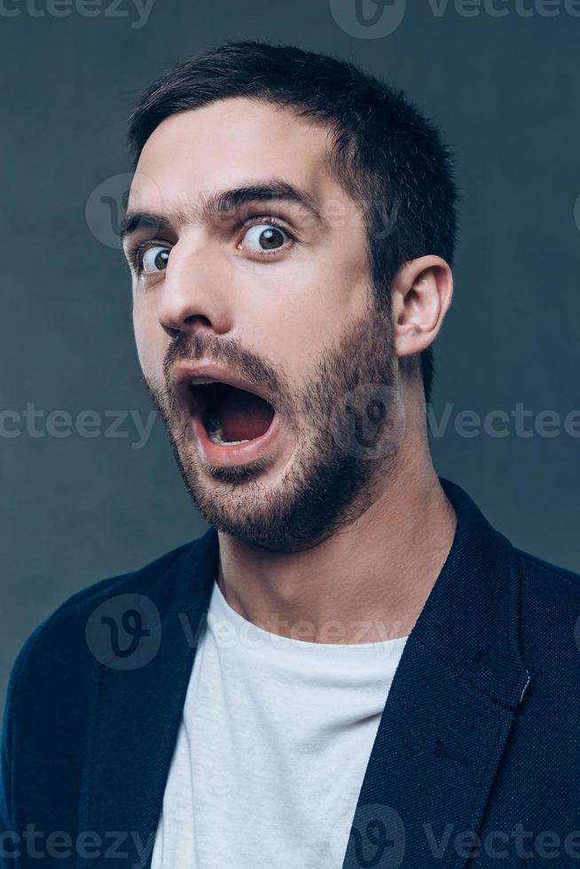 Stupid face. Portrait of grimacing young man opening his mouth and looking at camera while standing against grey background photo