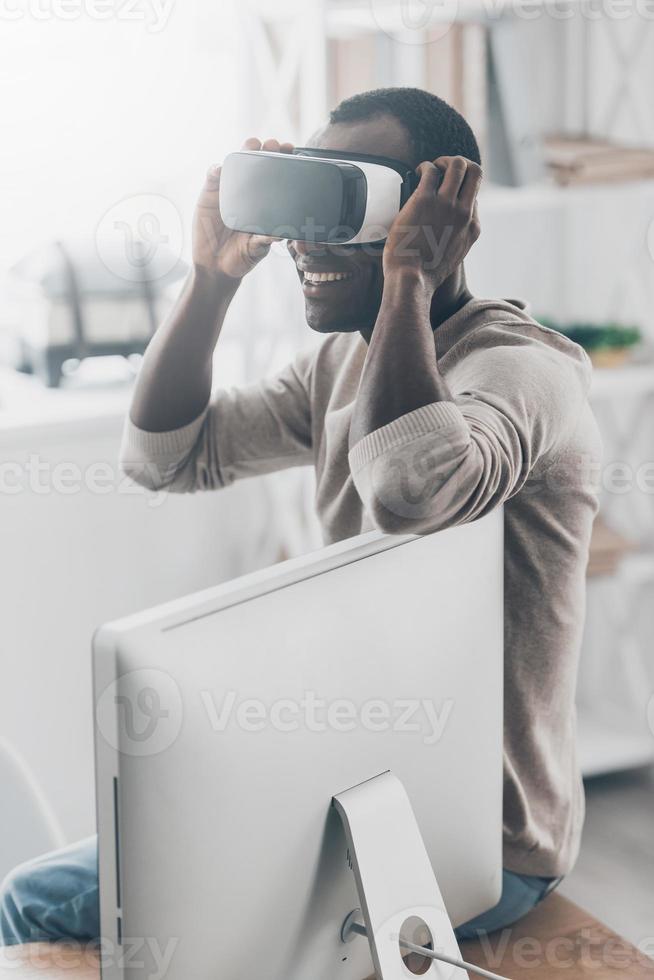 Everything is so real Handsome young African man adjusting his VR headset while sitting on the desk photo