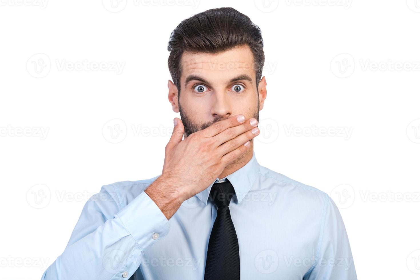 This is unbelievable Surprised young handsome man in shirt and tie covering mouth with hand and staring at camera while standing against white background photo