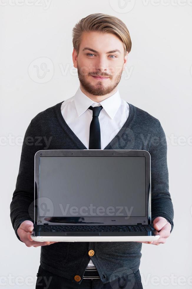 copie el espacio en el monitor. apuesto joven sosteniendo una computadora portátil y sonriendo mientras está de pie contra el fondo gris foto