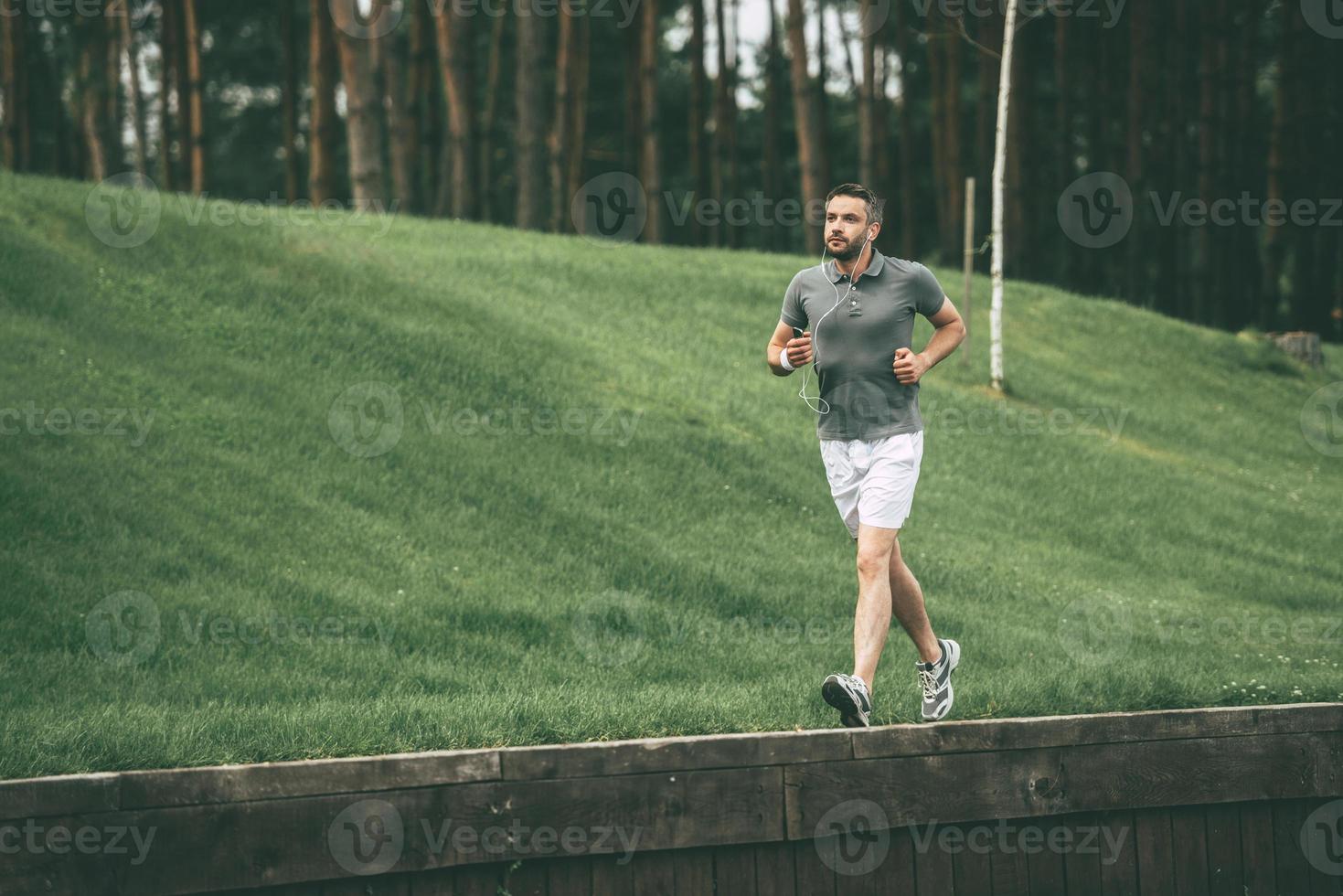 disfrutando del trote matutino. Longitud total de hombre confiado en ropa deportiva usando auriculares y corriendo en el parque foto