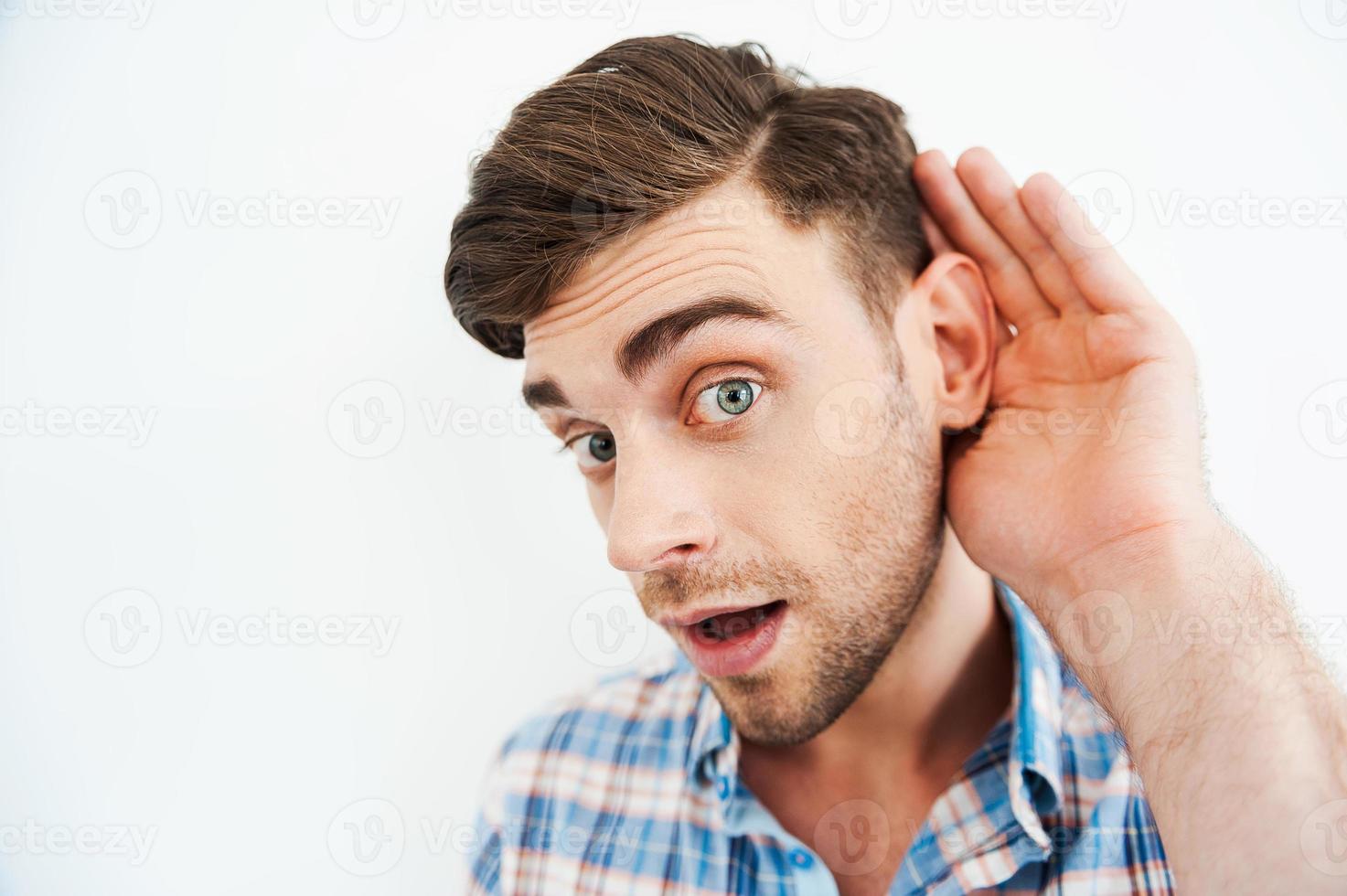 I want to know everything Curious young man holding hand behind ear and looking at camera while standing against white background photo